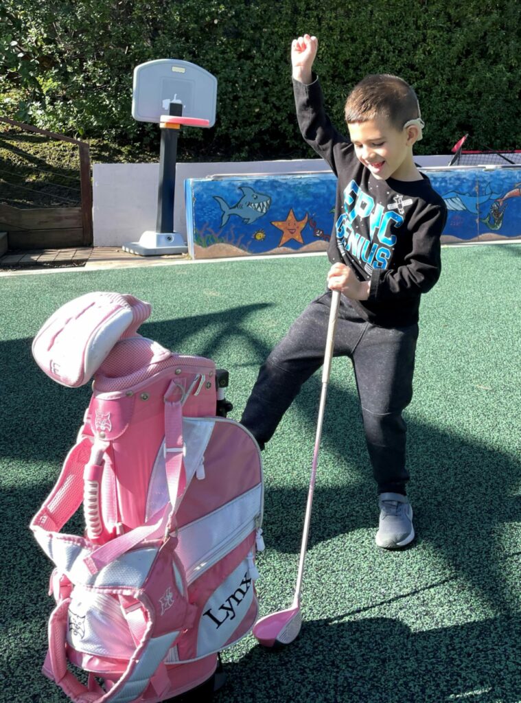 Child with hand in the air, golf club, and golf bag.